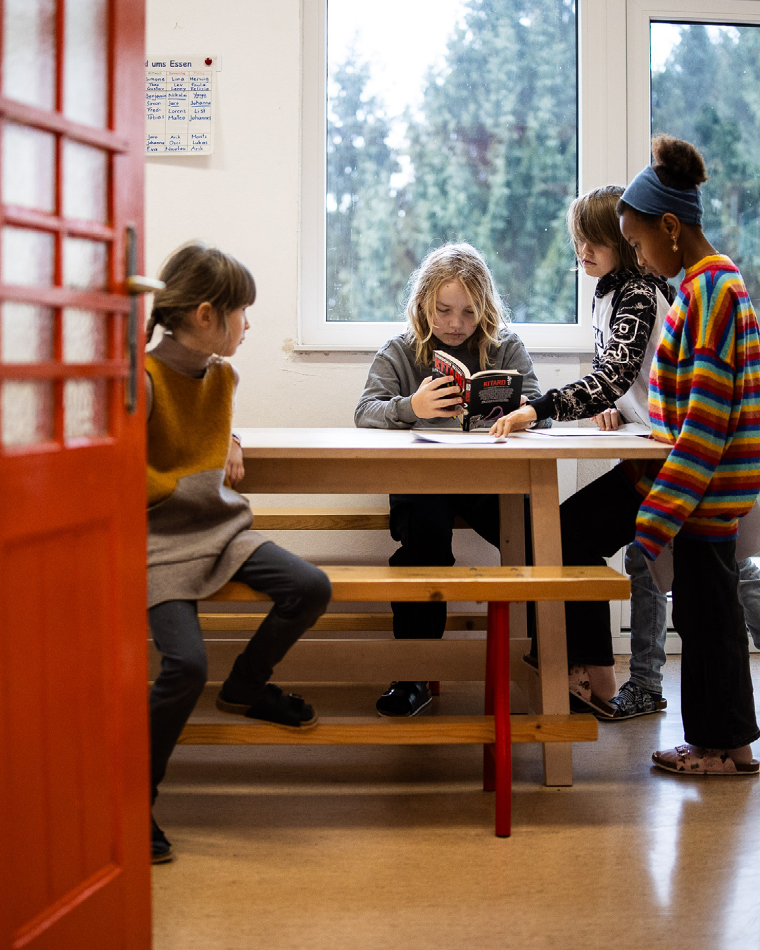 Vier Kinder lesen gemeinsam ein Buch an einem Tisch in einem Klassenraum; Fenster im Hintergrund, rote Tür links.