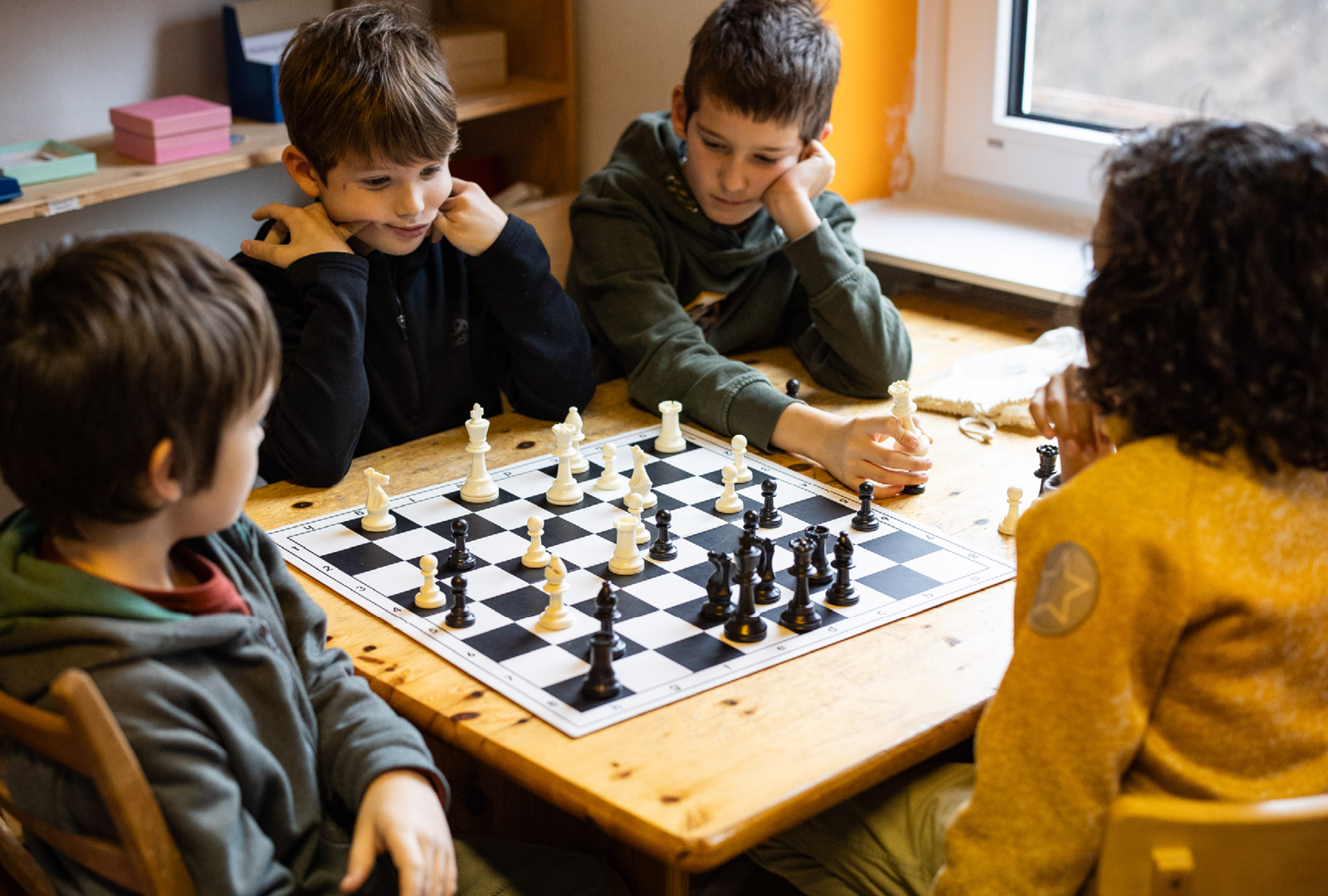 Vier Kinder spielen Schach an einem Holztisch in einem Lernraum; konzentrieren sich auf das Spiel, mit Schachfiguren auf dem Brett.