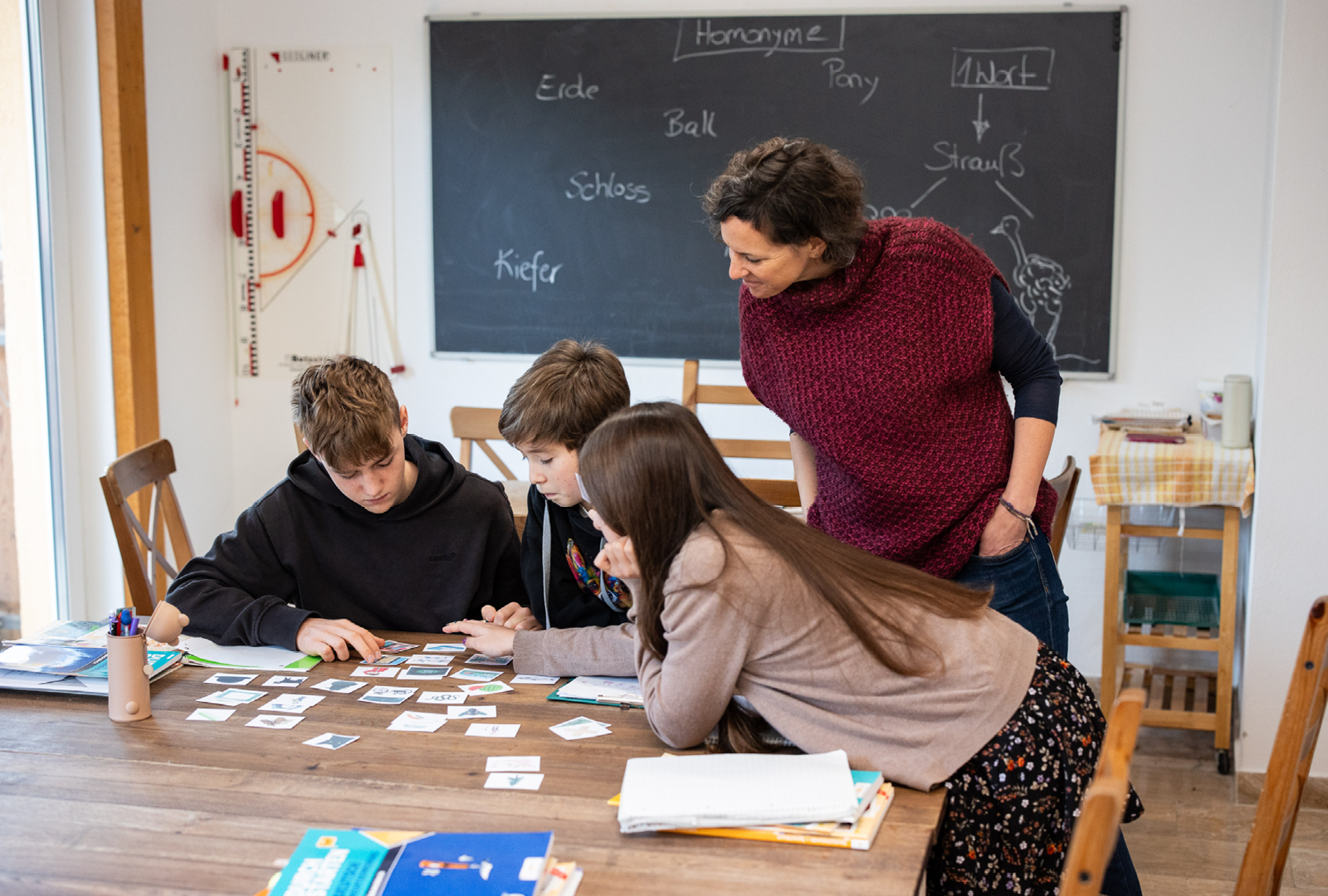 Lehrperson unterstützt drei Lernende beim Kartenlegen in einem Klassenzimmer; Tisch mit Karten, Tafel im Hintergrund.