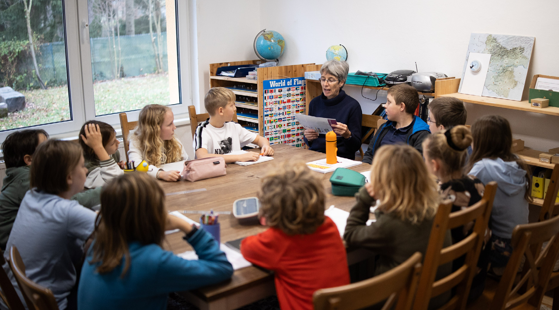 Lehrperson liest Grundschulkinder an einem Holztisch in einer Klasse etwas vor.