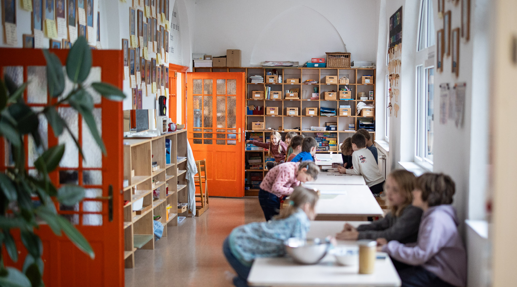 Klassenraum: Kinder arbeiten an Tischen, zeichnen; Regale und orange Tür im Hintergrund, Fensterzeile seitlich.