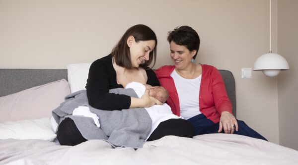 Zwei Frauen auf einem Bett; eine stillt ein Neugeborenes, die andere schaut lächelnd zu; Schlafzimmer mit grauem Kopfteil und Pendelleuchte.