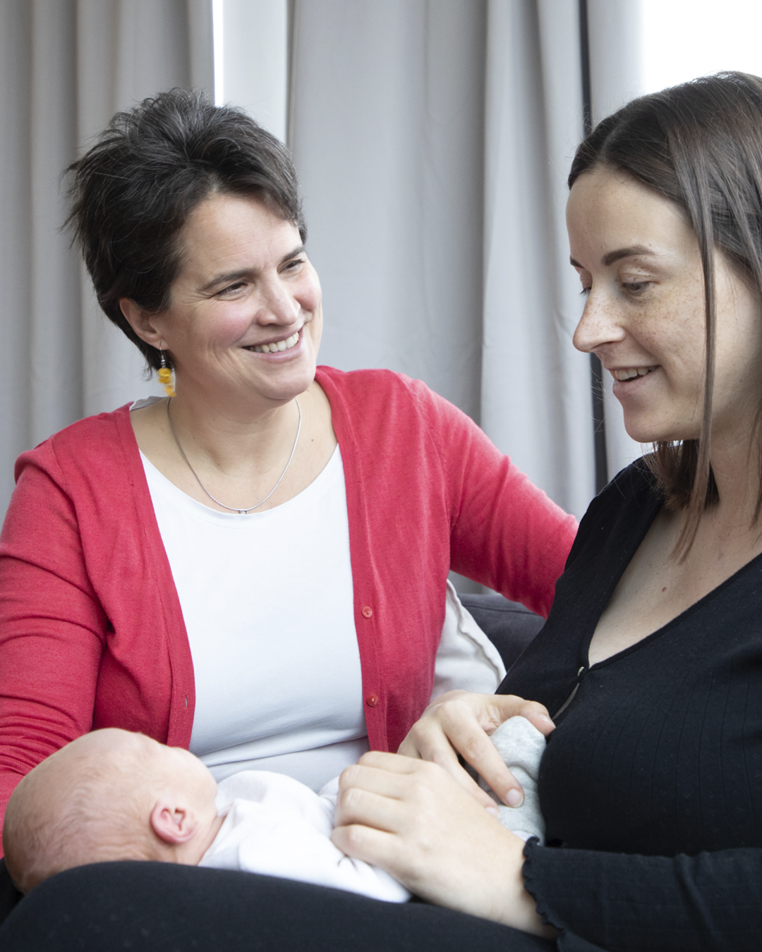 Zwei Frauen lachen und halten gemeinsam ein Neugeborenes; im Wohnzimmer mit Vorhängen.