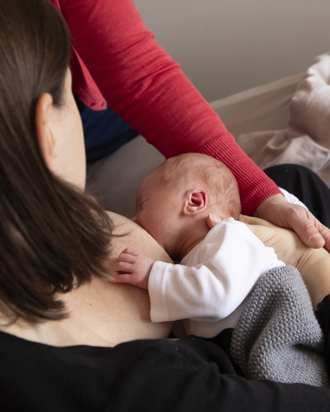 Baby wird an der Brust gestillt, während eine Person in rotem Pullover das Kind sanft hält; nah am Bett im Schlafzimmer.