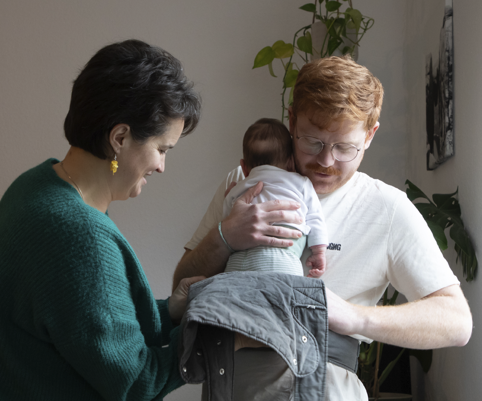 Säugling wird von einem Elternteil gehalten, der andere Elternteil lächelt daneben; Wohnzimmer mit Grünpflanze.