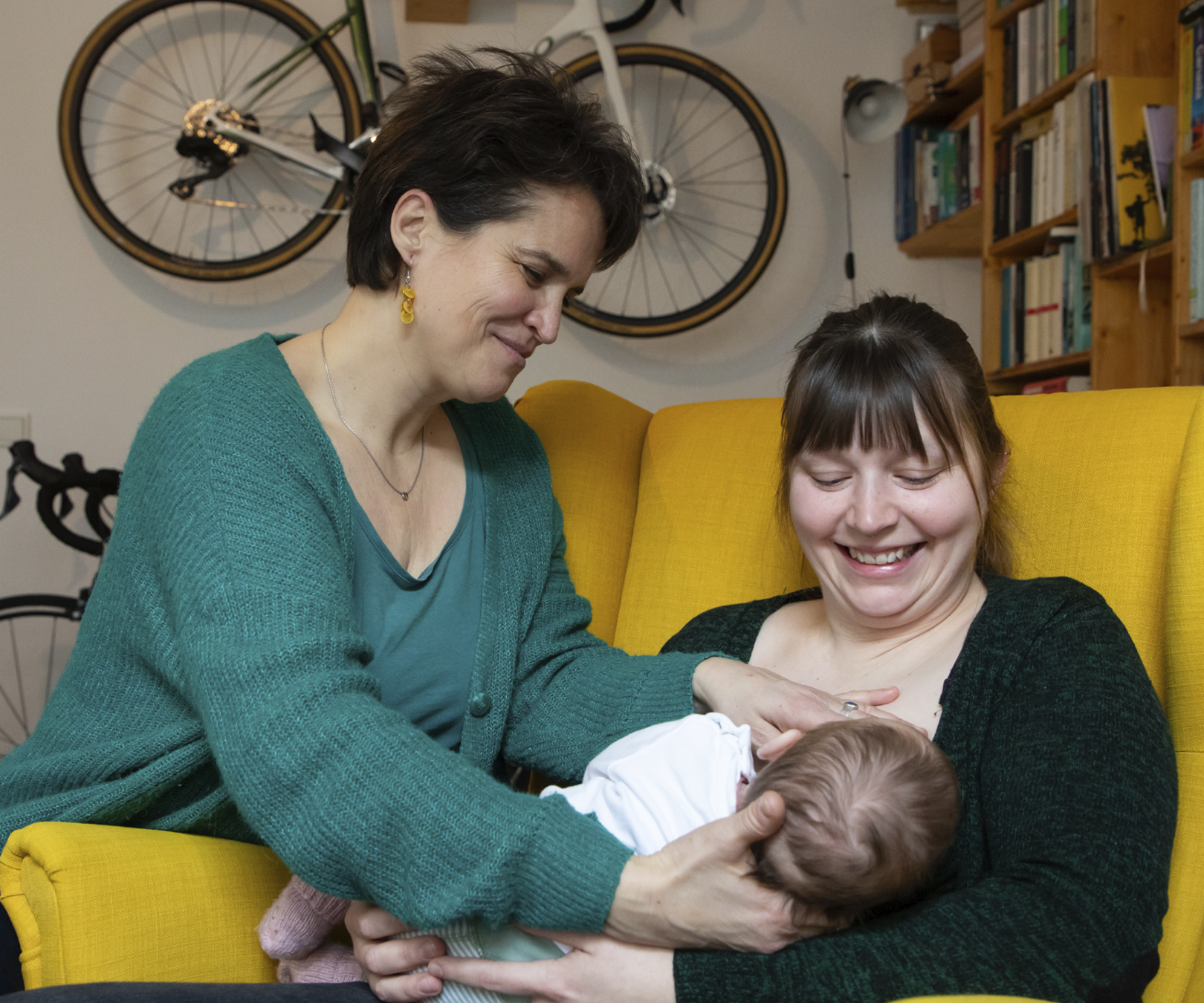 Baby in weißem Body wird von zwei Frauen im Wohnzimmer auf einem gelben Sessel gehalten; eine streichelt das Baby, die andere lächelt.
